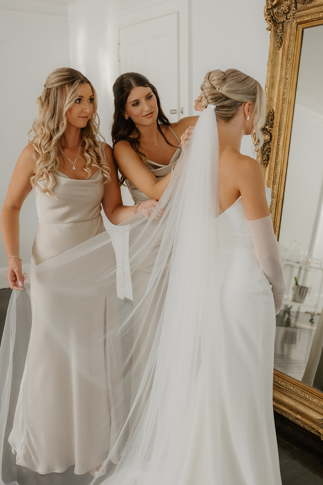 Bridesmaids help the bride adjust her long white veil in front of a gold mirror during prep.